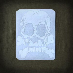 May include: White skull and crossbones design on a rectangular sticker. The skull has visible cracks and detailed features, with crossbones beneath. The sticker is on a dark green background, suitable for apparel or accessories.