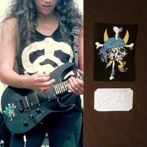 Puede incluir: Una guitarra eléctrica negra con un diseño de calavera y huesos cruzados, un parche negro con un diseño de calavera y huesos cruzados y una pegatina blanca con una firma. La persona lleva una camiseta sin mangas negra con un diseño blanco.