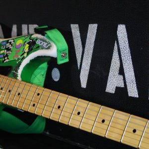 May include: An electric guitar in black and green, with a light wood neck and fretboard. The guitar has green details, stickers, and the word "BONES". The headstock displays the word "VAI" in white.