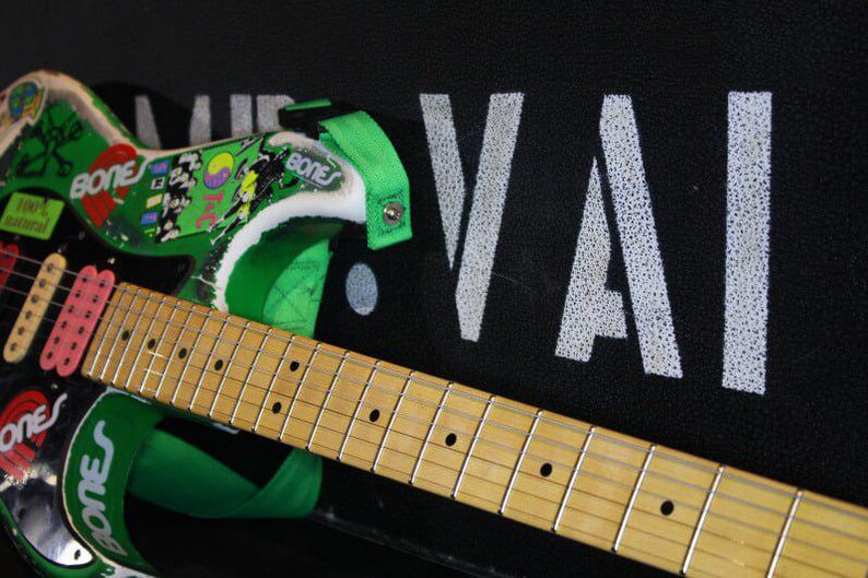 May include: An electric guitar in black and green, with a light wood neck and fretboard. The guitar has green details, stickers, and the word "BONES". The headstock displays the word "VAI" in white.