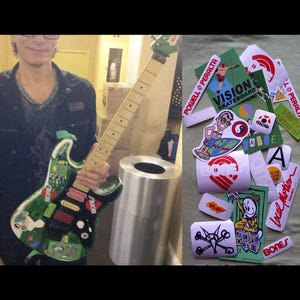 May include: An electric guitar with a green and black design, covered in stickers, held by a person. Stickers include "BONES", "POWELL PERALTA", and "VISION SKATEBOARD". A pile of stickers is also visible.
