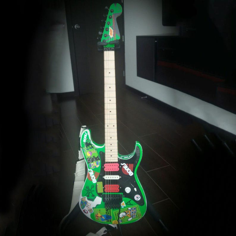 May include: A green electric guitar with a white neck and headstock. The body is decorated with stickers and the word "BONES". The guitar has red and black pickups and a black pickguard.
