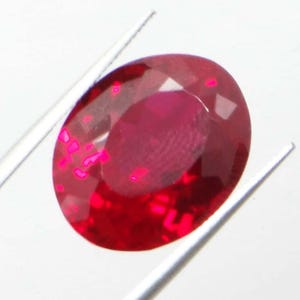 May include: A faceted, oval-shaped ruby gemstone, held by silver tweezers. The ruby is a deep red color with bright pink inclusions. The gem reflects light, showcasing its cut and clarity against a white background.