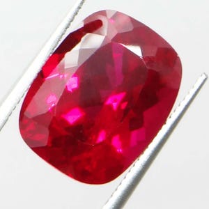 May include: A faceted, cushion-cut ruby gemstone held by silver tweezers. The ruby is a deep, saturated red with bright highlights. The gemstone's facets reflect light, creating a sparkling appearance. The background is a plain white.