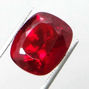 May include: A faceted, cushion-cut ruby gemstone held by silver tweezers. The ruby is a deep, saturated red color with a high degree of clarity, reflecting light. The gemstone's shape is a rounded square, showcasing its brilliance.