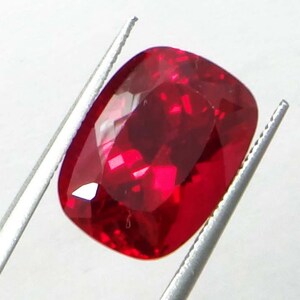May include: A faceted, cushion-cut ruby gemstone, held by silver tweezers. The ruby is a deep, saturated red color, with multiple facets reflecting light. The gemstone is a vibrant and eye-catching piece.