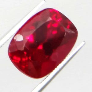 May include: A faceted, cushion-cut ruby gemstone, held by silver tweezers. The ruby is a deep, saturated red color with internal reflections. The gemstone is set against a white background.
