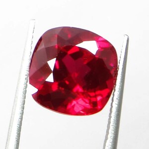 May include: A faceted, cushion-cut ruby gemstone held by silver tweezers. The ruby is a deep, saturated red color with bright internal reflections. The gemstone's facets catch the light, creating a sparkling effect.