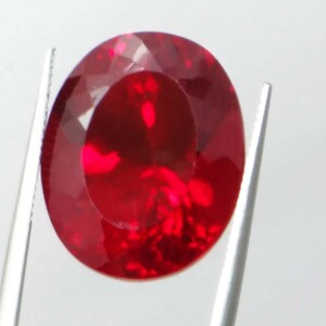 May include: A faceted, oval-shaped ruby gemstone held by silver tweezers. The gem is a deep, vibrant red color, with internal reflections creating a brilliant appearance. The tweezers are positioned on either side of the stone, showcasing its polished facets.