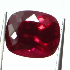 May include: A cushion-cut, deep red gemstone held by tweezers. The ruby exhibits multiple facets that reflect light, creating bright highlights. The gem's rich color and clarity are prominent against the white background.