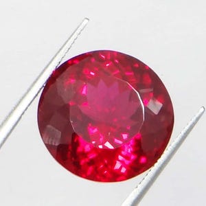 May include: A faceted, round-cut ruby gemstone, held by silver tweezers. The ruby is a deep, vibrant red color, with multiple facets reflecting light. The gemstone is the focal point, set against a plain white background.