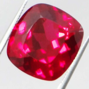 May include: A faceted, cushion-cut gemstone in a vibrant red hue. The gem is held by silver tweezers against a white background, showcasing its brilliant facets and internal reflections. The gemstone's color is a deep, saturated red.