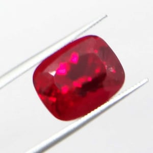 May include: A vibrant, cushion-cut ruby gemstone, held by silver tweezers. The ruby displays a deep red hue with internal light reflections. The gemstone's shape is a square with rounded corners, showcasing its facets.