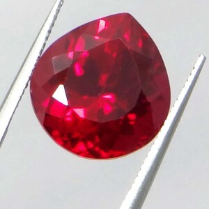 May include: A faceted, pear-shaped ruby gemstone. The gem is a deep, vibrant red color, held by silver tweezers against a white background. The cut of the stone creates multiple reflective surfaces.