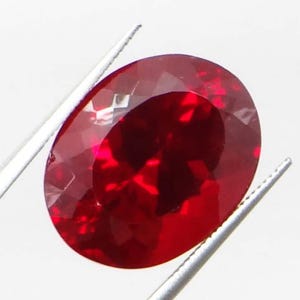 May include: A faceted, oval-shaped, deep red gemstone is held by silver tweezers against a white background. The gem's surface reflects light, showcasing its internal structure and rich color. This gemstone is likely a ruby, perfect for jewelry.