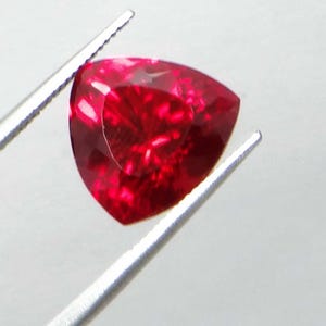 May include: A faceted, triangular-shaped, deep red gemstone is held by silver tweezers. The gem has a brilliant, reflective surface, showcasing internal patterns and light refraction. The gem's color is a rich, saturated red.
