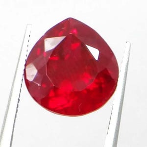 May include: A faceted, pear-shaped, deep red gemstone held by silver tweezers. The gem has multiple facets that reflect light, creating a brilliant sparkle. The gemstone is set against a white background.