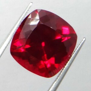 May include: A vibrant, cushion-cut ruby gemstone, held by silver tweezers. The gem's facets reflect light, showcasing its deep red color. The square shape with rounded corners is prominent.