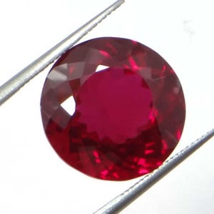 May include: A faceted, round-cut ruby gemstone, held by silver tweezers. The ruby is a deep, saturated red color, with multiple facets reflecting light. The gemstone is the focal point, showcasing its clarity and brilliance.