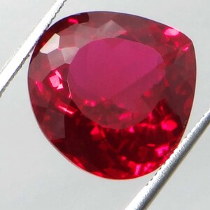 May include: A faceted, heart-shaped ruby gemstone. The gem is a deep, saturated red color, with multiple facets that reflect light. The ruby is held by silver tweezers, showcasing its brilliance and clarity. This gemstone is ideal for jewelry making.