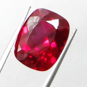 May include: A faceted, cushion-cut ruby gemstone, held by silver tweezers. The ruby displays a deep, saturated red color with internal reflections. The gem's facets catch the light, creating a brilliant display.