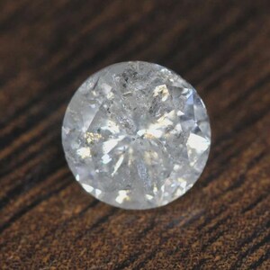 Può includere: Una gemma rotonda e trasparente con molteplici sfaccettature, che riflette la luce. La gemma ha un aspetto leggermente nuvoloso e si trova su una superficie di legno marrone scuro. La gemma è tagliata a brillante.