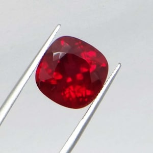 May include: A faceted, cushion-cut ruby gemstone, held by silver tweezers. The ruby is a deep, saturated red color with internal reflections. The gemstone's shape is square with rounded corners, and it has a high degree of clarity.
