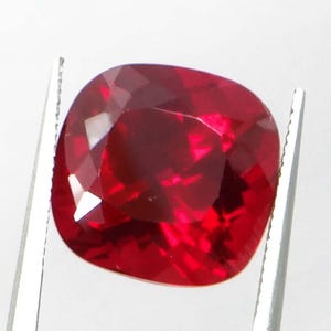 May include: A cushion-cut, faceted ruby gemstone, held by silver tweezers. The ruby is a deep, saturated red color, with multiple facets reflecting light. The gemstone is square with rounded corners.