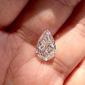 Könnte beinhalten: Ein birnenförmiger Diamant mit brillanten Facetten, gehalten in einer Hand. Der farblose Edelstein funkelt und reflektiert das Licht. Der Schliff des Diamanten ist darauf ausgelegt, seine Brillanz und sein Feuer zu maximieren und seine Klarheit und Schönheit zu zeigen.