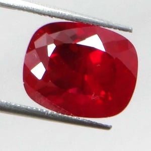 May include: A cushion-cut, faceted ruby gemstone held by tweezers. The ruby is a deep, saturated red color with bright internal reflections. The gemstone's facets catch the light, creating a sparkling effect. The tweezers are silver.