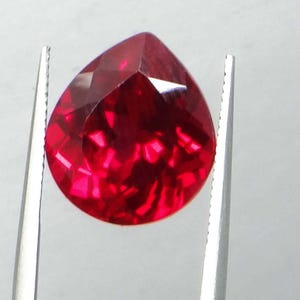 May include: A faceted, pear-shaped, deep red gemstone is held by silver tweezers against a white background. The gem's cut reflects light, creating bright highlights and shadows. The gemstone is a vibrant, saturated red.