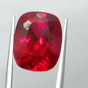 May include: A faceted, cushion-cut ruby gemstone, held by silver tweezers. The ruby is a deep, saturated red color with multiple facets reflecting light. The gemstone's shape is rectangular with rounded corners.