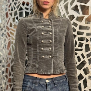 May include: A grey military-style jacket with a high collar and multiple silver buttons. The jacket has a fitted silhouette and is made of a soft, velvety material. The jacket is paired with blue denim jeans.
