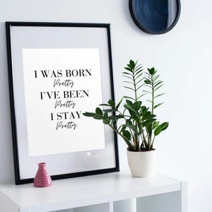 Könnte beinhalten: Ein gerahmter Druck mit dem Text "I WAS BORN Pretty, I'VE BEEN Pretty, I STAY Pretty" in einem schwarzen Rahmen. Eine kleine rosa Vase und eine Topfpflanze stehen auf einem weißen Regal. Ein dunkles kreisförmiges Objekt hängt an der weißen Wand.