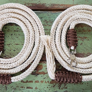 May include: Two cream-coloured rope dog leads, coiled and arranged side-by-side. Each lead has a brown woven handle and a silver-coloured clasp. The leads are on a distressed, green-painted wooden surface.