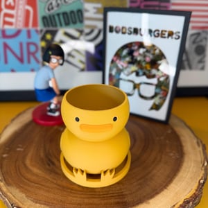 May include: A yellow duck-shaped planter with a smiling face, set on a wooden slice. The planter has a round body and a base with claw-like feet. In the background, a framed print reads "BOBSBURGERS" and a figurine of a character.
