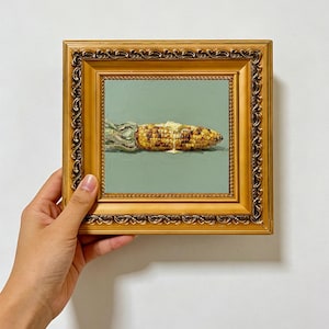 May include: An oil painting of a single ear of grilled corn with melting butter, set against a solid sage green background. The artwork is encased in an ornate, metallic gold frame with intricate embossed detailing along the edges.