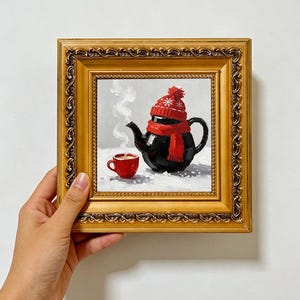 Könnte beinhalten: Ein Ölgemälde in einem dekorativen Goldrahmen zeigt eine schwarze Teekanne mit einer roten Strickmütze und einem Schal, neben einer kleinen roten Tasse mit aufsteigendem Dampf. Der Hintergrund ist hellgrau mit weißen, schneeartigen Akzenten.