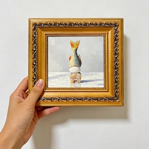 May include: An oil painting in a decorative gold frame depicting a fish tail pointing upwards, balanced on a white egg, which rests on a slice of fish. The composition features a light grey background and a white surface with subtle shadows.