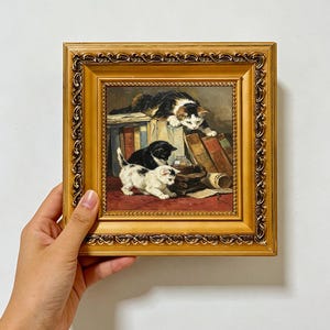 以下が含まれることがあります： 装飾が施された金色のフレームに入った正方形の油絵。本の山と小さなガラス瓶と戯れる3匹の子猫が描かれています。1匹の子猫は本の上に座り、他の2匹は赤い表面にいます。テキストはSALARCOLLECTIONSと書かれています。