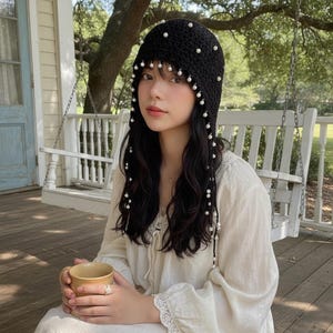 May include: A black crocheted hat adorned with white pearls. The hat features dangling pearl strands and is worn by a person. The person is wearing a cream-colored dress and holding a cup. The background includes a porch swing and trees.