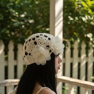 May include: A cream-colored crochet hat with a floral accent. The hat features an open weave design and is adorned with two white fabric flowers. The hat is worn by a person with long, dark hair.