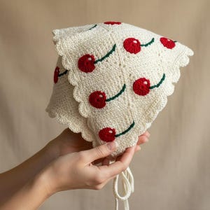 Op de afbeelding: Crèmekleurige gehaakte hoed met een geschulpte rand, versierd met rode kersenapplicaties en groene stelen. De hoed heeft een striksluiting. Een handgemaakt, grillig accessoire.