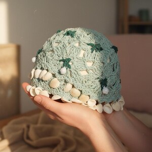 May include: A light blue crocheted hat with a seashell and bead trim. The hat features a textured, open weave design with dark green leaf accents and small white beads. The hat is a unique, handmade accessory.