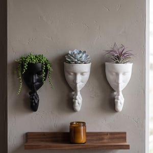 May include: Three wall-mounted planters shaped like human faces with a finger held to the lips in a shushing gesture. The planters are black, speckled gray, and white, each holding a different green plant, positioned above a wooden shelf.