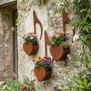 Może przedstawiać: Trzy rdzewiejące metalowe donice w kształcie nut wiszą na kamiennej ścianie. Każda donica ma pojemnik w kształcie serca wypełniony kolorowymi kwiatami. Ściana pokryta jest zielenią.