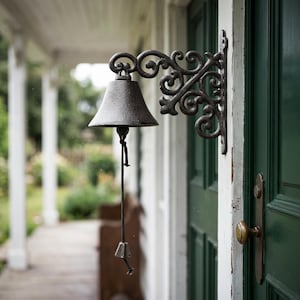 May include: A cast iron bell with an ornate bracket, mounted on a green door. The bell is dark gray with a rope pull and a small clapper. The bracket features scrolling designs. The background shows a porch and a glimpse of a garden.