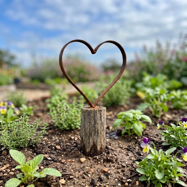 May include: A rustic metal heart sculpture, mounted on a weathered wooden post, stands in a garden setting. The heart is made of dark brown metal. The background features green plants and a blue sky.