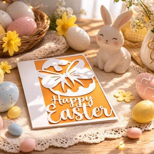 Puede incluir: Una naturaleza muerta con temática de Pascua con un conejito de cerámica blanco, huevos decorados y una tarjeta que dice "Felices Pascuas". Los huevos son de varios colores y diseños, con una cesta de huevos en el fondo.