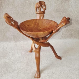 May include: A brown wooden bowl supported by three carved legs, each featuring an animal figure at the top. The central bowl is smooth and circular, while the legs are intricately shaped with detailed carvings of an elephant and other animals.